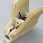 Close-up of a wooden clip with visible grain, showcasing its metal spring mechanism. The clip is partially open and rests on a light, neutral background.