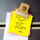 A wooden clip with Food 2 Go engraved holds colorful sticky notes on a surface. The yellow note reads: Dont Forget! Call school @ 9am. Zoom Meeting @ 12:30pm. -Dad-.