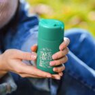 A person holding a green reusable bottle with the text Take Hikes and a mountain design. The background shows blurred green grass and a person wearing jeans.