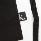 Close-up of a black fabric with a small black tag featuring a lowercase white letter k and a period. The fabric has straight edges and a loose string or strap drapes beside it on a white background.