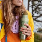 A person wearing a yellow raincoat and orange sweater holds a green water bottle with botanical designs. They are outdoors with trees and water visible in the background.