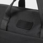 Close-up of a black fabric bag with two handles. The material has a textured weave, and there is a small rectangular black patch on the side. The lighting highlights the texture and stitching details.