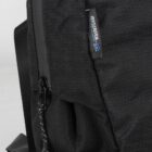 Close-up of a black backpack featuring the Waste2Wear logo in white and blue on a small tag. The fabric has a textured pattern, and a sturdy zipper is visible at the side.