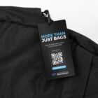 Close-up of a black fabric with a tag that reads, More Than Just Bags. Out of the Waste Cycle & Into Your Gear. Includes a QR code and website link to waste2gear.org. The tag is attached with a string.