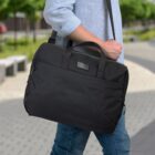 A person in a light blue shirt and jeans carries a black laptop bag on their shoulder. The background shows a blurred outdoor path lined with trees.