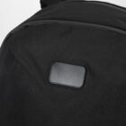 Close-up of a black backpack with a small rectangular patch on the front. The fabric has a subtle textured pattern, and the patch appears to be made of leather or a similar material.