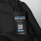 Close-up of a black tag attached to a black fabric. The tag reads More Than Just Bags with a QR code below. It includes the website waste2gear.org and the tagline Out of the waste cycle & into your gear.