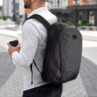 A man in a white shirt carries a black backpack and a takeaway coffee cup, walking along a paved, modern urban street with gray buildings and green shrubs in the background.