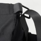 Close-up of a black fabric strap looped through a metal ring on a bag. The fabric has a textured pattern, and the hardware has a matte finish. The background is white, highlighting the details of the strap and ring connection.