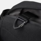 Close-up of a black fabric bag with a textured surface, showing a zipper partially open. A black strap is visible, passing through a triangular metal loop, which is secured by a metal clip.