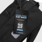 A close-up of a black fabric bag with a tag attached. The tag reads More Than Just Bags and features a QR code, the website waste2gear.org, and the phrase Out of the waste cycle & into your gear.