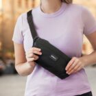 A person wearing a light purple t-shirt holds a black crossbody bag labeled STMNT across their chest. The background is a blurred urban outdoor setting with warm lighting.