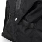 Close-up of a black backpack pocket featuring a button closure and a zippered compartment above, made from durable fabric. The texture and stitching details are visible, highlighting the bags craftsmanship and design.