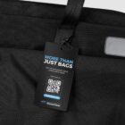 A black bag with a tag attached. The tag reads More Than Just Bags and includes a QR code. Below it is the text Out of the waste cycle & into your gear and a website link waste2gear.org.