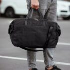 A person in gray jeans holds a large black duffel bag with both hands while standing in a parking lot. A white car is blurred in the background.