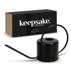 A sleek black watering can with a long spout and curved handle is placed in front of a black and brown box labeled keepsake collection.