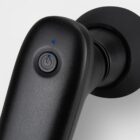 Close-up of a black massager with a power button and two small blue indicator lights on its handle. The massager has a smooth, sleek design and a circular attachment at one end, set against a light gray background.