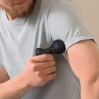 A bearded person uses a black massage gun on their upper arm. They are wearing a light gray t-shirt. The background is a neutral color.