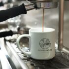 A white mug with a Coffee House logo is placed under an espresso machine spout, with coffee being freshly brewed into it. The kitchen equipment is visible in the background, giving a warm and inviting atmosphere.
