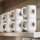 White mugs with Coffee House Premium Aroma logos are neatly stacked on a shelf. The stack has three columns, each with three mugs, set against a tiled background. The mugs feature a design with a coffee cup emblem.