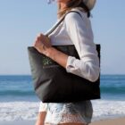 A woman in a sun hat and white blouse carries a black tote bag with a green tree logo and the word EcoHome on it. She stands on a beach with waves in the background.