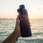 A hand holds a dark blue water bottle against the backdrop of a sunset over the ocean. The sun is partially visible, casting a warm glow on the water and bottle. A bracelet is worn on the wrist of the hand holding the bottle.