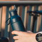 A hand holding a dark blue reusable water bottle with a flip lid and loop handle. A blurred background shows vertical railing bars. A smartwatch is worn on the wrist of the hand holding the bottle.