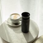 A black travel mug and a cup of cappuccino with a spoon rest on a round white table. The table is near a window, and sunlight softly illuminates the scene, casting shadows on the surface.