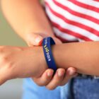Person wearing a blue wristband with KIDS ART GALA printed on it. They have a striped red and white shirt and jeans, adjusting the wristband on their arm.