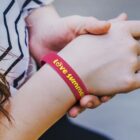A person wearing a red wristband with the words love summer in yellow. The wrist is held with the other hand, and the person is wearing a white shirt with black stripes.