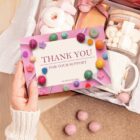 A hand holding a colorful Thank You for Your Support card surrounded by candies. The card is part of a gift package with sweets, a mug, and a jar of marshmallows, set on a textured surface.