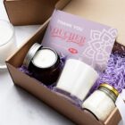 A gift box containing a candle in a brown jar, a white candle in a glass, a jar with a cork lid, and a spa and beauty voucher worth $150. The box is lined with purple shredded paper.
