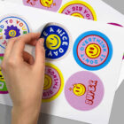 A hand peeling a circular sticker with a smiley face and text Have a Nice Day. Other visible stickers say Don’t Overthink It, Super, and Be Kind to Yourself, all featuring colorful backgrounds and designs.