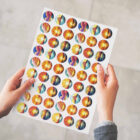 Two hands are holding a sheet of circular stickers. The stickers feature vibrant illustrations of a landscape with a sunset or sunrise, mountains, and a stylized sun. The background is a light-colored surface.