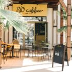 Outdoor seating area of a coffee shop named RIO coffee with tables and chairs. A sign reads We Are OPEN near the entrance. The area is shaded by palm trees, creating a welcoming atmosphere.
