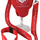 A wine glass is held in a red fabric holder with a neck strap. The strap is printed with Swan Hill Region Food & Wine Festival in white text. The holder is designed to be worn around the neck for convenience.