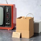 A retro-style orange TV is on a table next to a brown gift box with a white handle visible inside. A beige card with text about a special edition is leaning against the box. A blurred object is in the background.