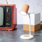 A small, modern desk lamp with a cork-textured shade stands on a gray surface. Next to it is a vintage red television. The background is minimal with a subtle gray tone.