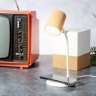 A vintage red TV stands beside a modern white lamp with a cylindrical shade. Below the lamp, a smartphone is wirelessly charging on a circular base. The background features a potted fern and a white and brown box.