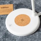 A white wireless charging pad with a cork center rests on a gray stone surface. A small card is partially visible in the background.