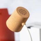 Close-up of a modern desk lamp with a cylindrical cork shade and a white neck. The lamp is shown against a blurred background featuring hints of red and white objects.