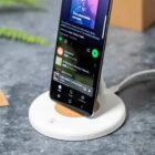 A smartphone displaying a music streaming app is positioned upright on a white wireless charging stand. The phone screen shows a playlist. The setup rests on a gray stone surface, with a blurred green plant in the background.