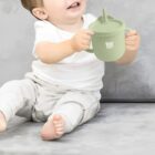 A toddler wearing a white shirt and light-colored pants sits on a gray surface, holding a green sippy cup labeled Baby Kids. The child appears to be smiling, and the cup has a straw lid.