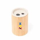 A cylindrical wooden pencil sharpener with two holes and a light-colored top. It features a colorful logo with the letters ZAM in blue, yellow, and red on the front.