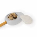 A wooden handle attached to a gray oval tool with a circular opening, containing ashes and small debris. A round, flat, light-colored lid is next to it. The tool and lid are set against a white background.