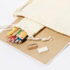An open beige drawstring bag spills colorful pencils, a wooden ruler, a sharpener, and an eraser onto a brown notebook with a white edge, set against a plain white background.