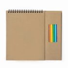 A brown spiral notebook with a cardboard cover is next to a set of six colored pencils in a matching cardboard box, arranged in rainbow order: red, orange, yellow, green, blue, and purple.