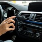 A person in a cars drivers seat is attaching a smartphone to a dashboard-mounted holder. The dashboard displays various controls and an air vent. The persons hand is holding the phone near the holder.