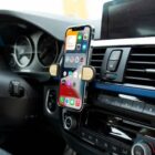 A smartphone with a colorful screen is mounted on a cars air vent using a black and wood-finish holder. The phone displays various app icons. The cars dashboard and control buttons are partially visible in the background.