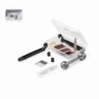 A bicycle repair kit displayed on a white background. It includes tire levers, patches, a metal multi-tool, rubber solution in a tube, sandpaper, and small cylinder caps. The items are arranged around an open plastic box.